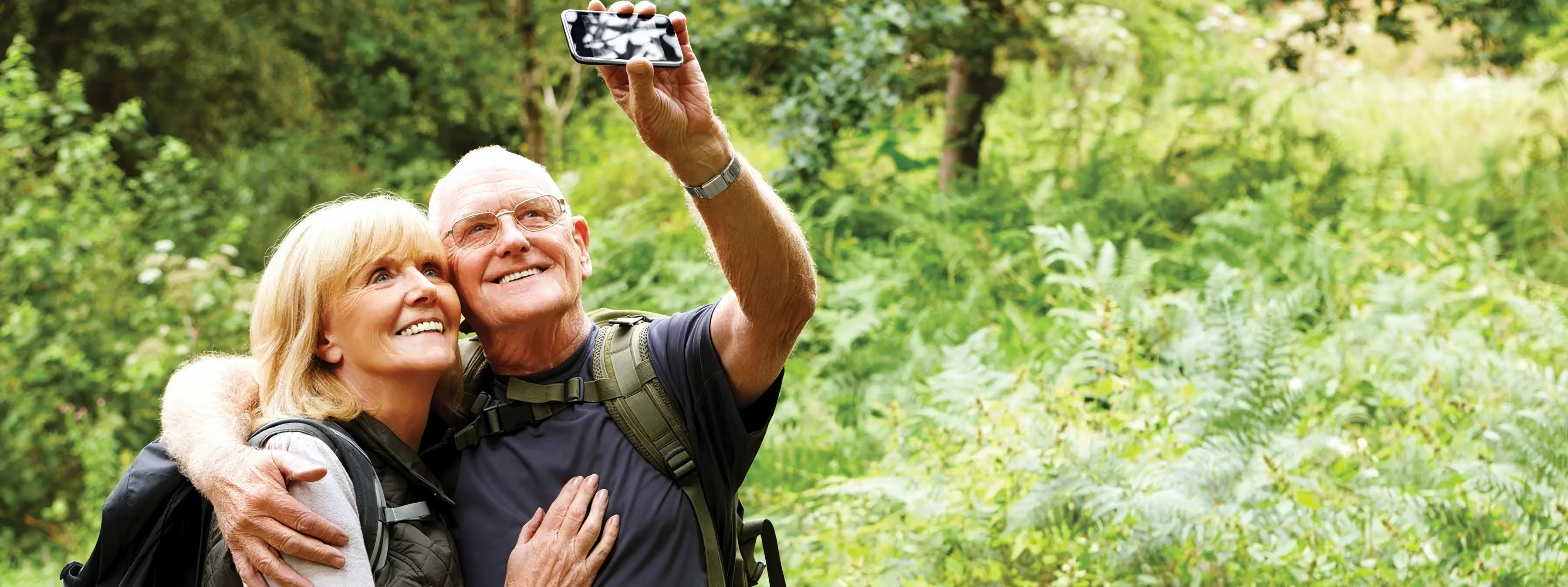An older couple takes a selfie smiling together outdoors amidst greenery.