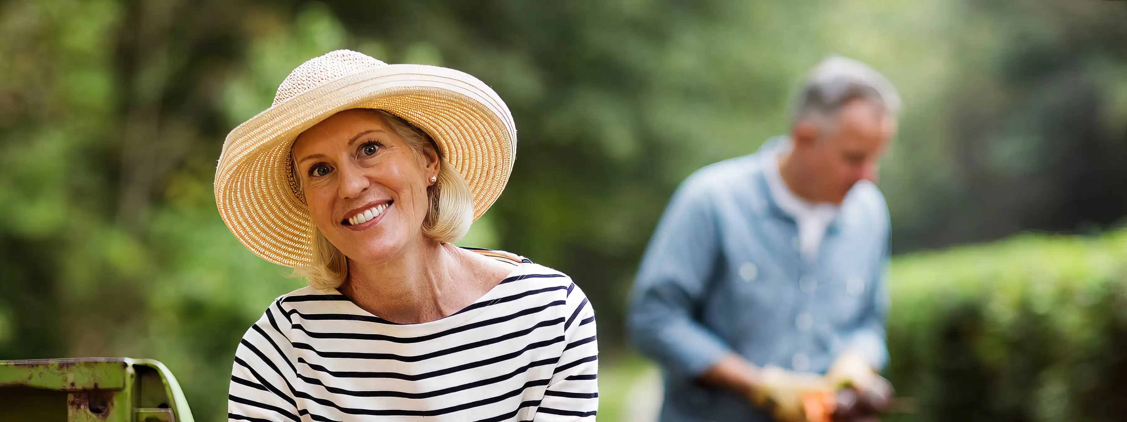 A woman in a straw hat smiles while gardening, with a man working in the background.