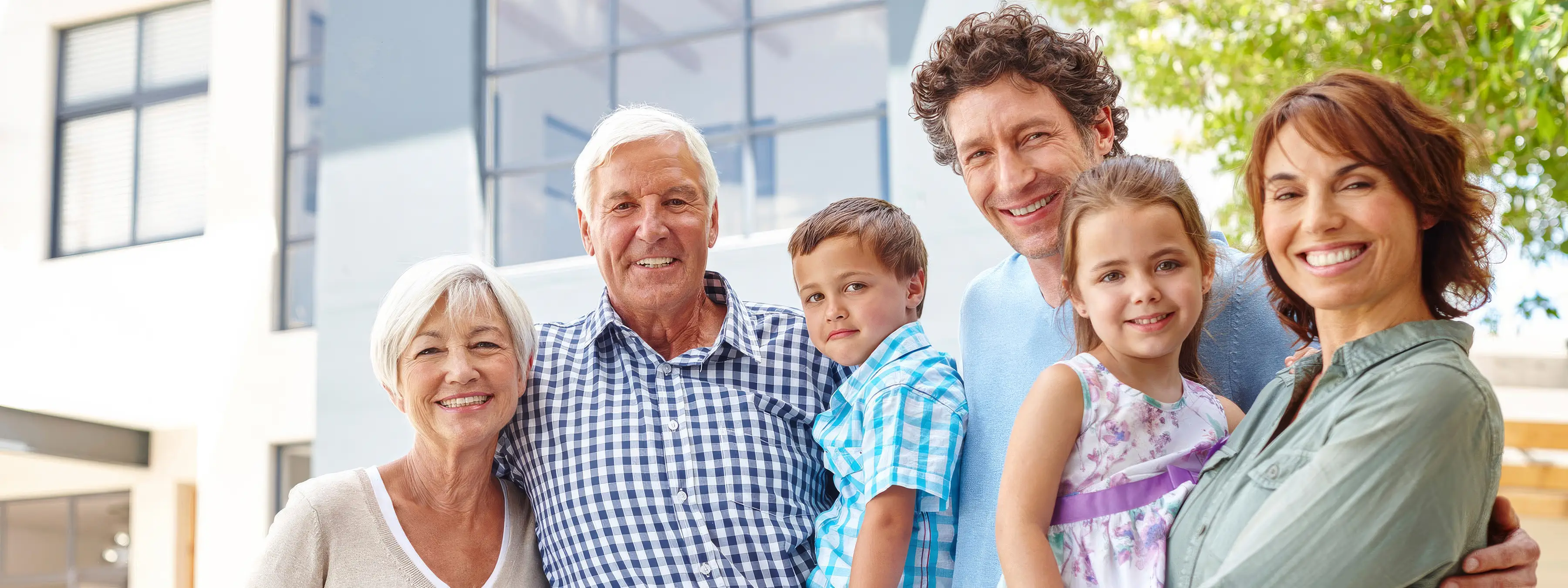 A multi-generational family group smiling together outdoors, standing close and posing for a photo.