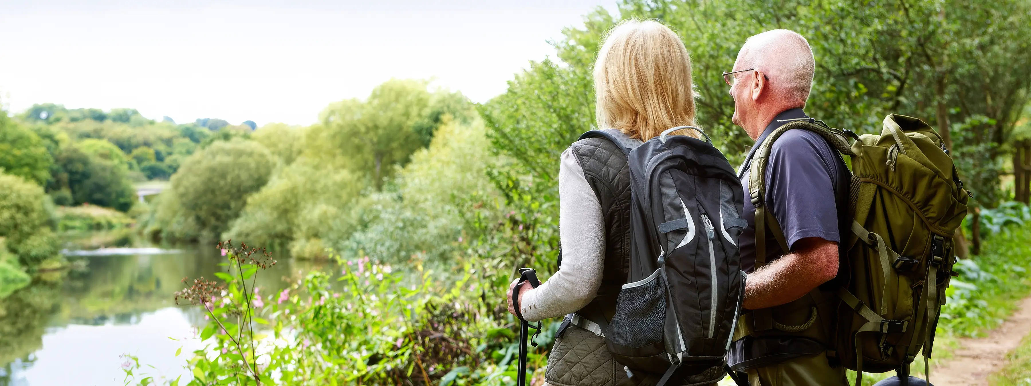 A man and woman with backpacks stand by a river, looking at the lush landscape and trees around them while hiking.