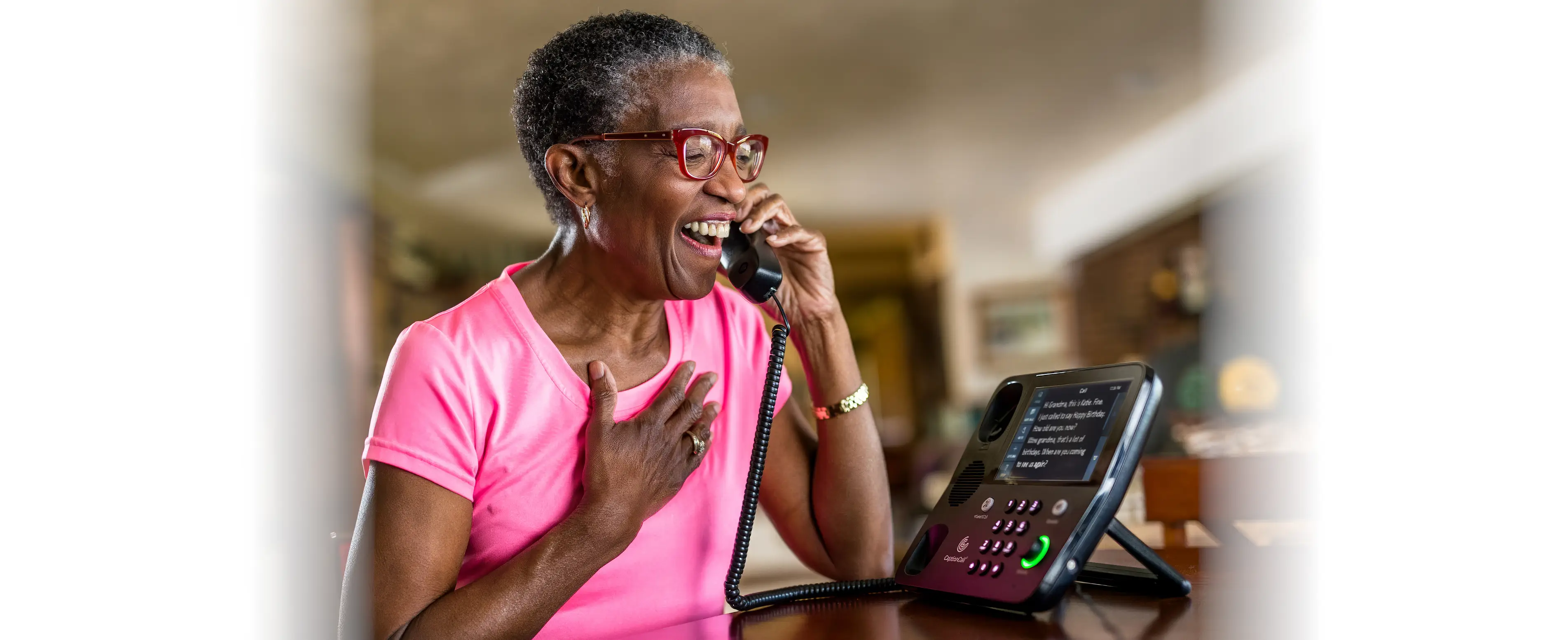 A senior woman in a pink shirt laughs while using a landline phone, appearing joyful during a conversation.