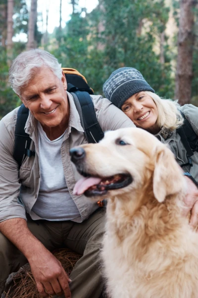 A couple smiles while posing with their golden retriever during a hike in a forested area.