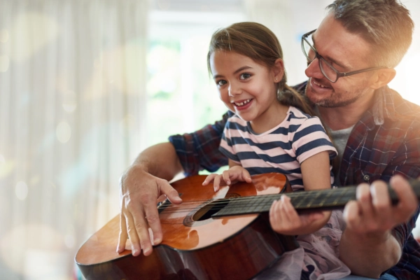 A man plays guitar while a young girl smiles and strums the instrument together with him.