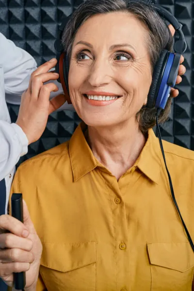 A woman in a yellow shirt wearing headphones smiles as a person adjusts them, inside a hearing test booth.